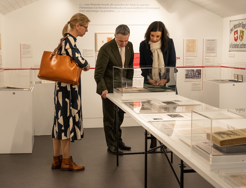 Drei Personen betrachten gemeinsam Exponate in einer Vitrinen-Ausstellung, umgeben von Dokumenten und Informationstafeln an den Wänden. Die Szene spielt in einem modern gestalteten Museumsraum.