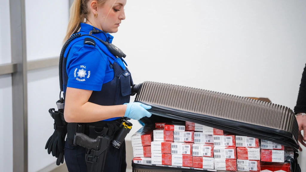 A FOCBS officer opens a suitcase filled with cigarette packs. She is wearing a uniform and carefully inspects the contents. The scene likely takes place at a customs checkpoint.