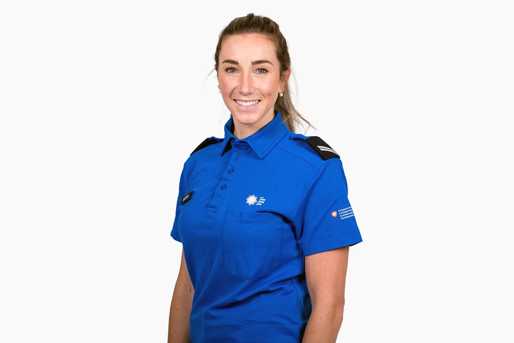 A woman in a blue uniform smiles at the camera. She is wearing a shirt with badges on the shoulders.