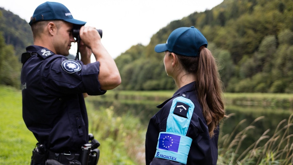 Two Frontex officers monitoring a border in a wooded area. One uses binoculars while the other observes attentively.  