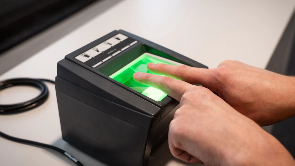 A person places two fingers on a fingerprint scanner that glows green. The device is on a table and connected by a cable. The scene represents a biometric identification.
