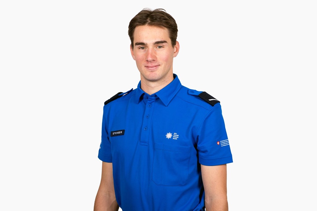 A young man in a blue customs uniform stands against a white background. Badges and a name tag are visible on his uniform.