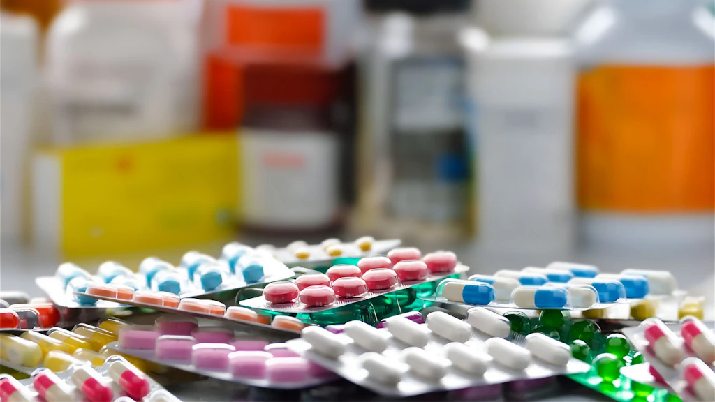 Packaged medicines in different sizes, colours and shapes lie in a jumble on a table. In the background, other medicines in cardboard boxes and bottles can be vaguely recognised.