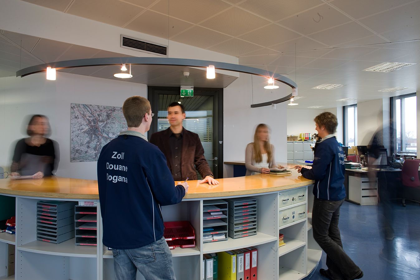 Two customs officers stand at a reception desk and speak with visitors. Other people and office desks are visible in the background.