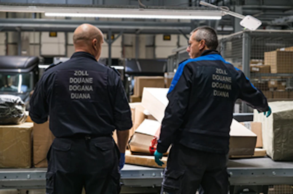 Two FOCBS employees stand at a conveyor belt with parcels, inspecting shipments. They wear uniforms with "Zoll Douane Dogana Duana" written on the back. The scene takes place in a warehouse or logistics center.