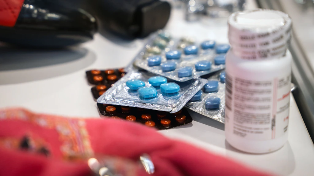Various blister packs with blue and brown tablets and a white medicine bottle are lying on a table. A red cloth is blurred in the foreground, with other objects in the background. The scene suggests a medication inspection.