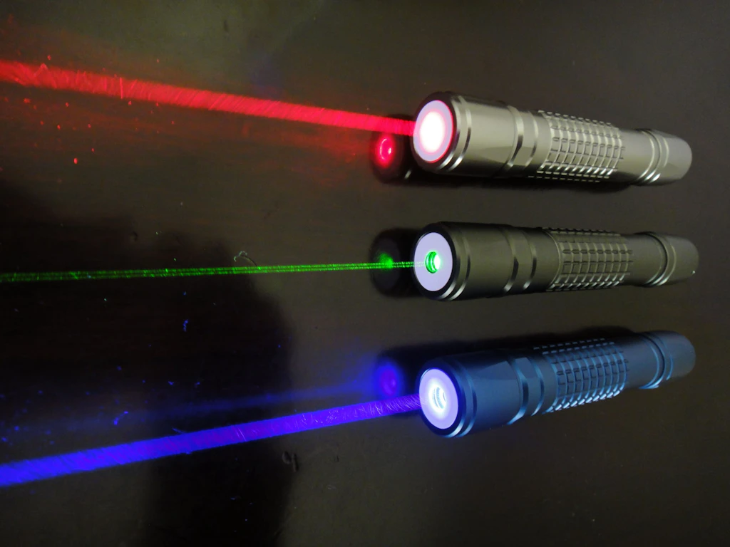 Three switched-on laser pointers are lying side by side on a dark surface. The laser beams are clearly visible in red, green, and blue. Each pointer emits a distinct light beam in its respective color.