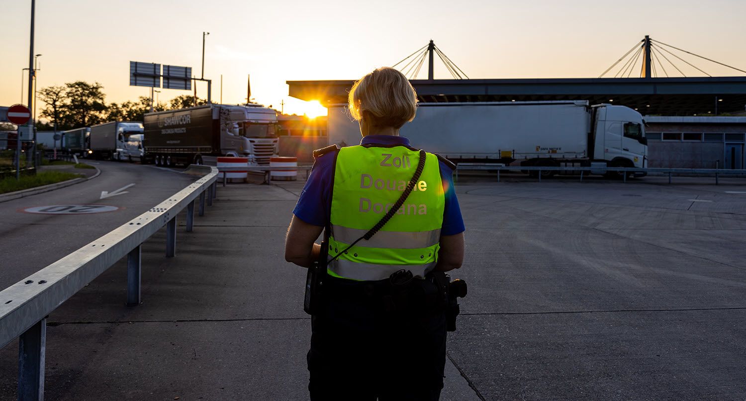 A person in a customs vest stands at sunrise in a parking lot, with trucks parked in the background.  