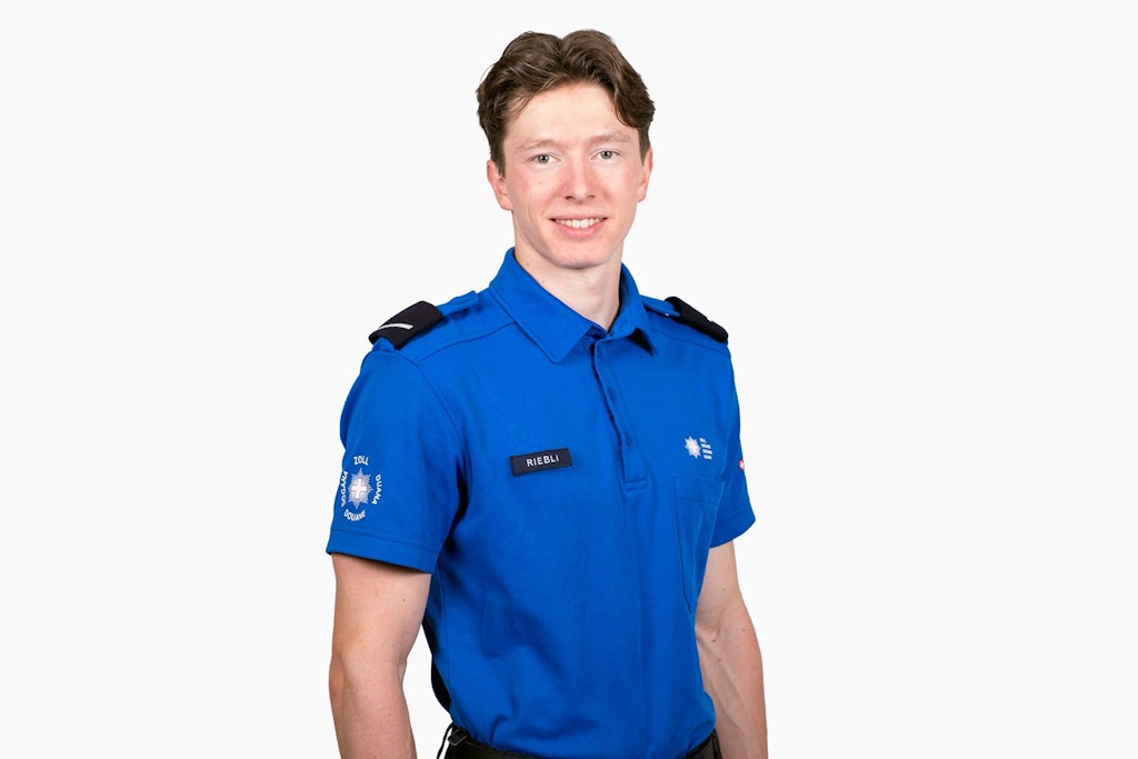 An employee of the FOCBS in a blue uniform with a badge stands in front of a neutral background. He wears a name tag and has a friendly demeanour.