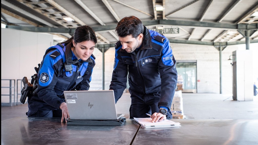 Two employees of the FOCBS working together on a laptop in a warehouse. They are wearing uniforms and appear focused.