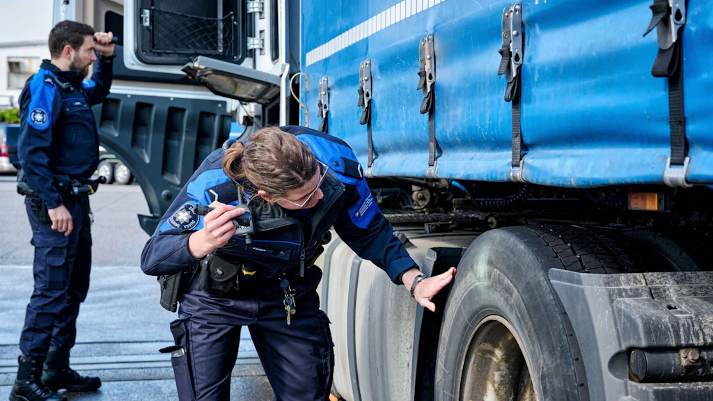 Two employees of the FOCBS inspect a truck. One person carefully checks the area around the wheel, while the other stands by the cab. Both are in uniform and appear focused.