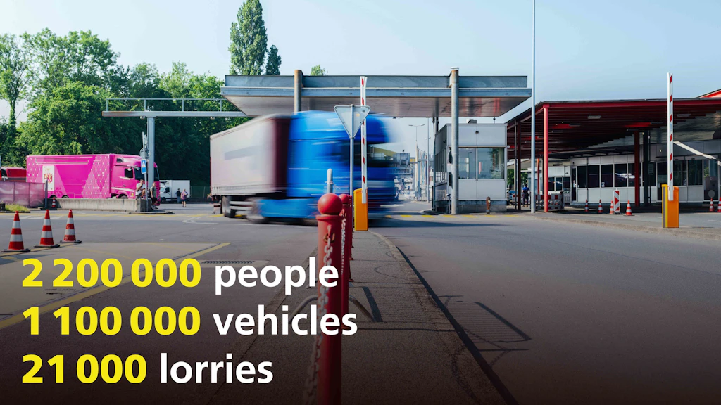 A border crossing with multiple lanes, where a blue truck is passing through. In the background, a pink truck is visible and traffic cones are placed on the left. In the foreground, statistical figures about people, vehicles, and trucks are displayed.