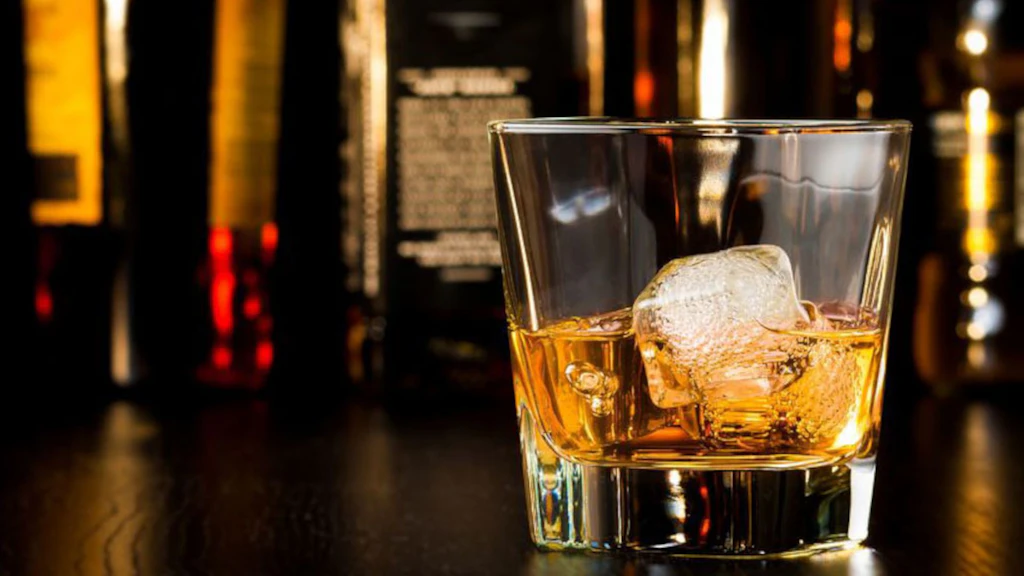 Un verre rempli au tiers d'un liquide brun clair et de deux glaçons est posé sur une table brun foncé. En arrière-plan, on distingue des silhouettes de bouteilles.
