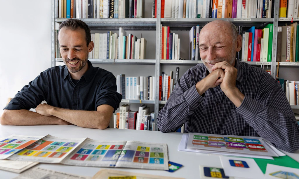 Deux hommes sont assis a une table dans une bibliotheque et examinent des projets historiques de vignettes autoroutières etalees devant eux. Tous deux sourient et semblent concentres sur les vignettes colorees et les documents.  