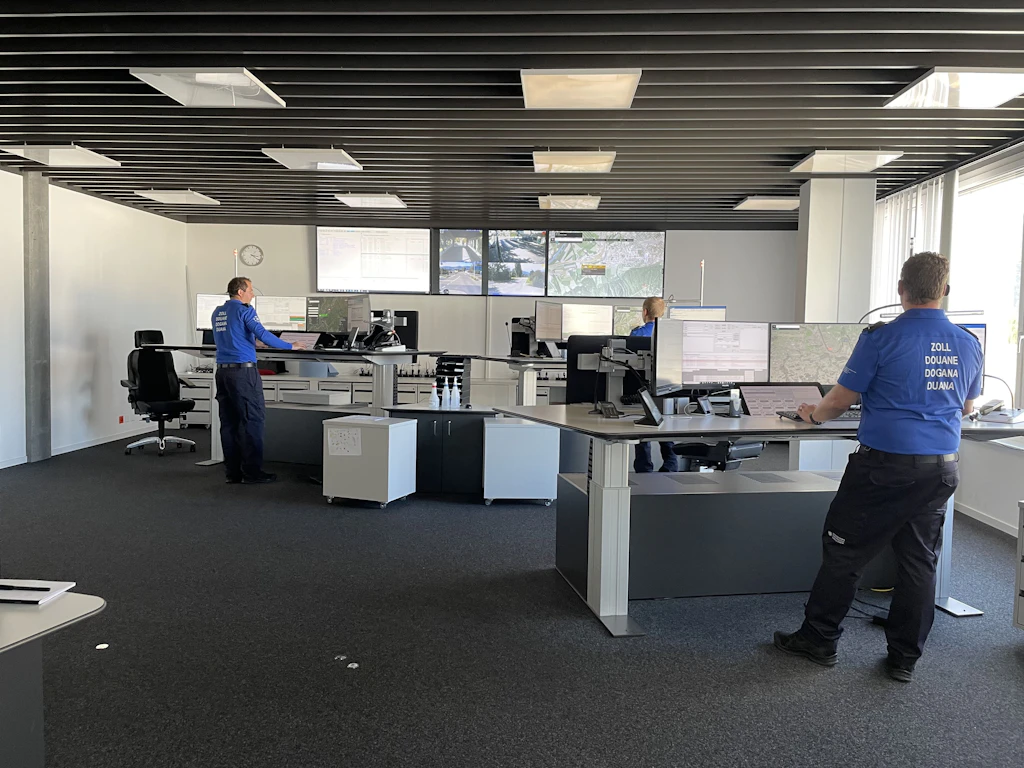 Plusieurs collaborateurs de la douane en uniforme bleu travaillent dans la centrale d engagement Est, dans un grand local moderne equipe de nombreux ecrans. De grands moniteurs muraux affichent des cartes et des images video de la region frontaliere, tandis que la lumiere du jour entre par une large baie vitree.  