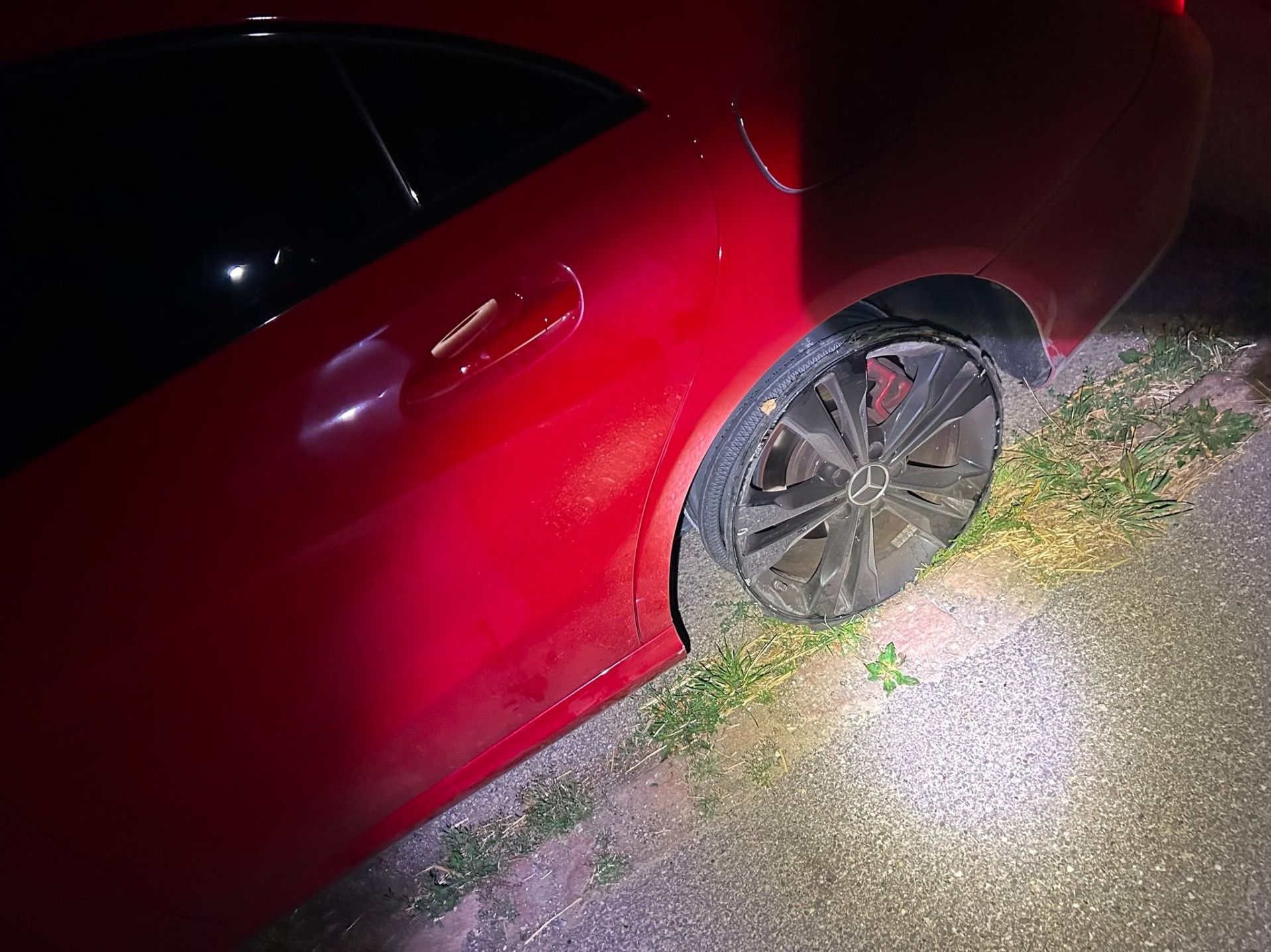 Une voiture rouge est garée au bord de la route, le pneu arrière est complètement à plat et endommagé. Le véhicule semble avoir roulé malgré ce dommage. La scène est éclairée de nuit à la lampe torche.