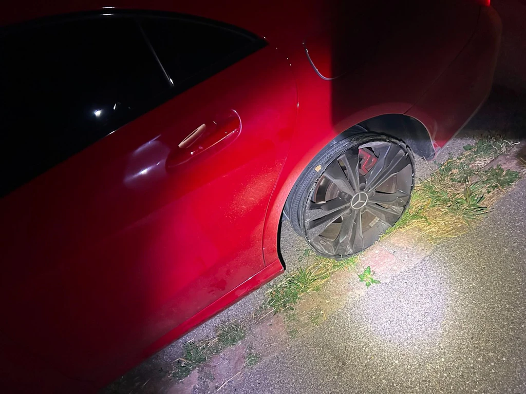 Une voiture rouge est garée au bord de la route, le pneu arrière est complètement à plat et endommagé. Le véhicule semble avoir roulé malgré ce dommage. La scène est éclairée de nuit à la lampe torche.