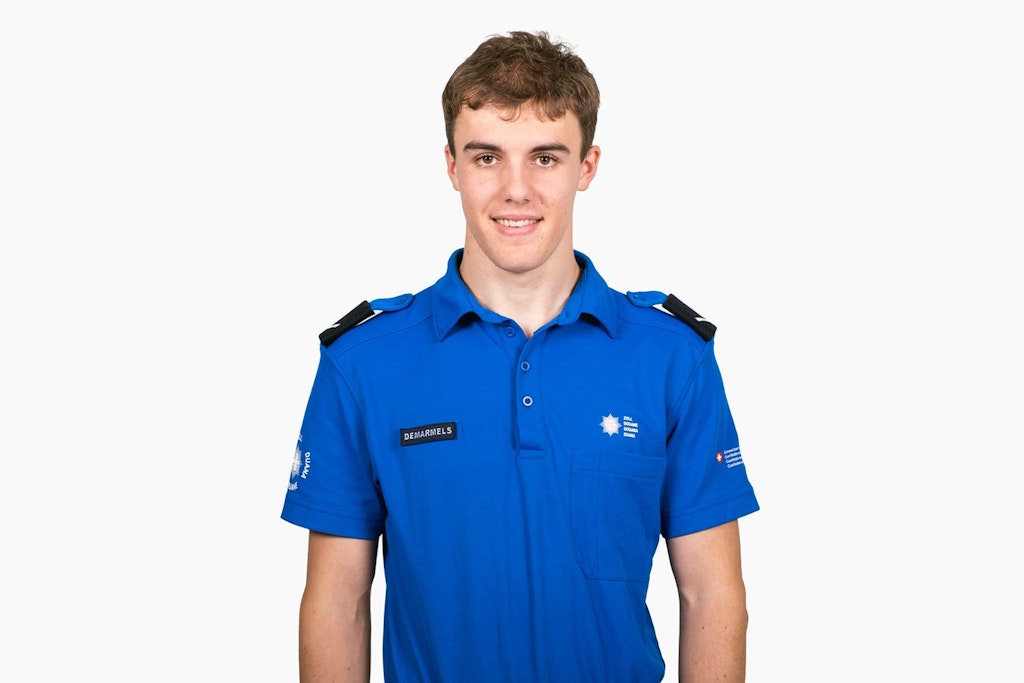 Un jeune homme en uniforme bleu de douane se tient devant un fond blanc. Divers insignes sont visibles sur l'uniforme.