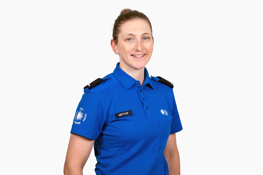 Une femme en uniforme bleu avec l'inscription "Douane" se tient devant un fond blanc. Elle sourit et porte un badge avec le nom "Mettler".