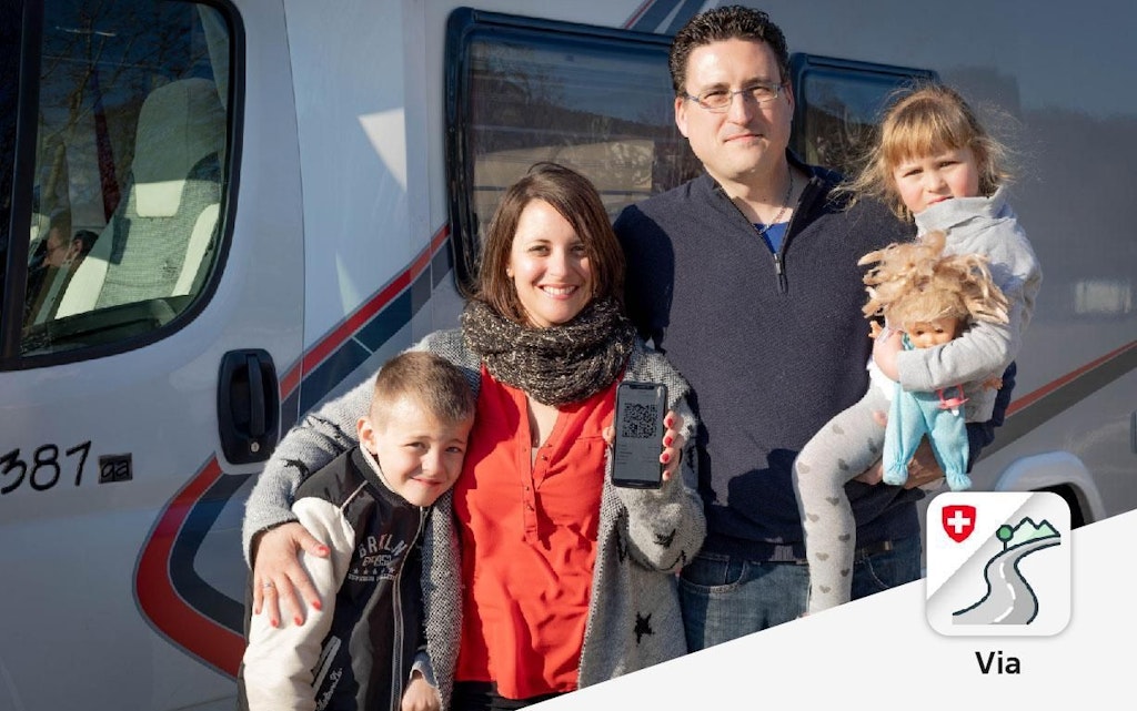 Une famille se tient devant un camping-car. La mère tient un smartphone avec l'application VIA. Un enfant est dans les bras du père.