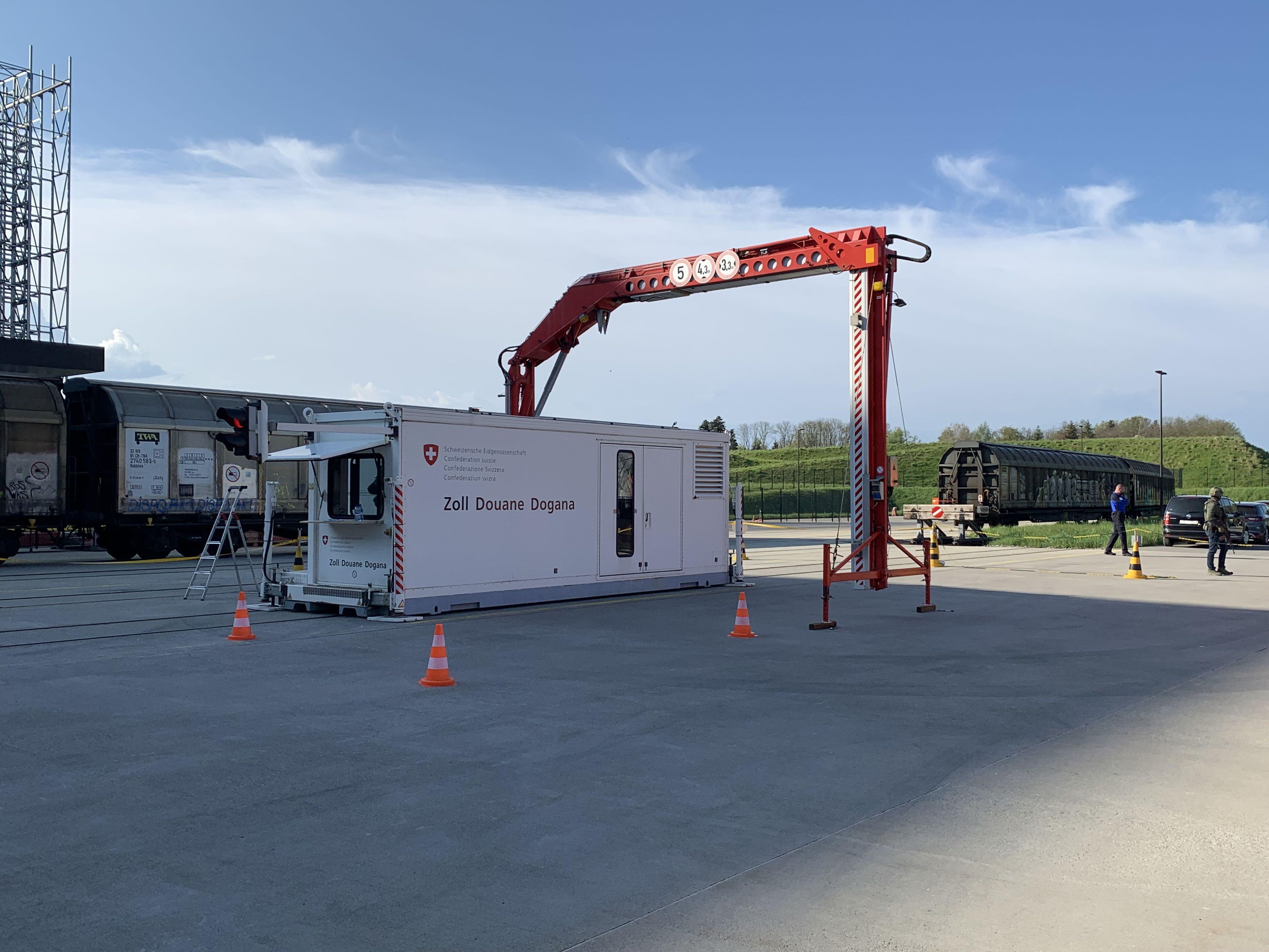 Une unité mobile de contrôle douanier est située sur un quai à côté d'un train de marchandises. L'unité est blanche avec des marques rouges, entourée de cônes de circulation sous un ciel clair.