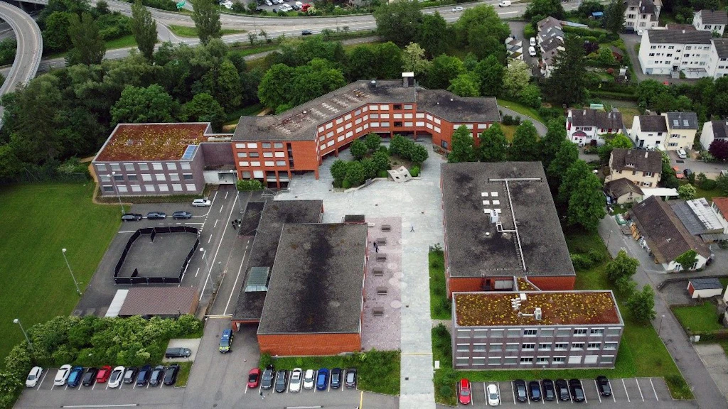L’image montre une vue aérienne d’un complexe moderne composé de plusieurs bâtiments à toit plat, de parkings et d’espaces verts. À l’arrière-plan, on aperçoit des maisons et des routes. Les bâtiments sont entourés d’arbres et de routes.