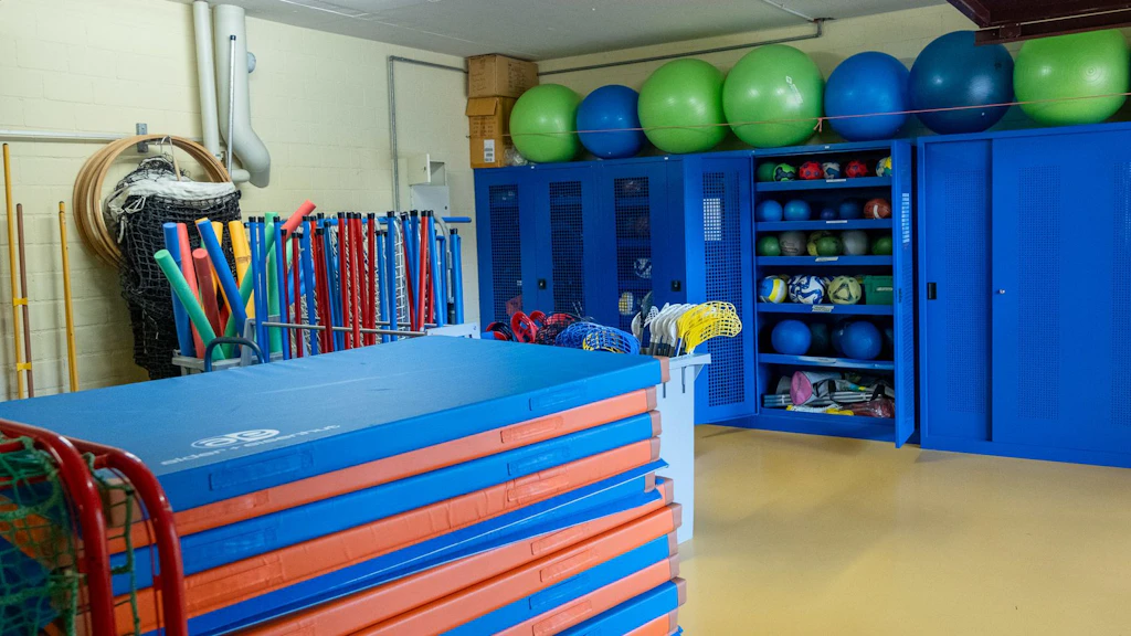 Une salle de matériel sportif bien organisée avec des armoires bleues, des ballons de gymnastique colorés, des tapis, des cerceaux et divers équipements de sport. Le matériel est soigneusement rangé et prêt à être utilisé pour les cours de sport.