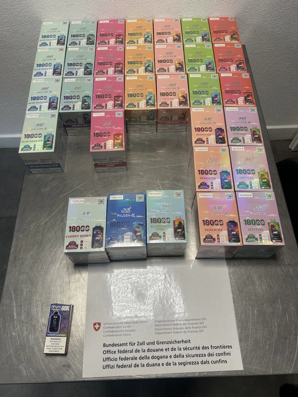 Sur une table se trouvent de nombreux paquets de cigarettes électroniques jetables de différentes saveurs, triés et exposés. Au premier plan, une pancarte de l’Office fédéral de la douane et de la sécurité des frontières est visible. Les produits sont classés par couleur et présentent différentes marques et variétés.  