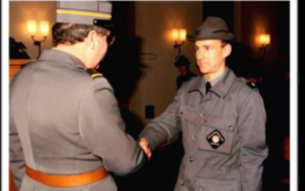 Deux hommes en uniforme se serrent la main dans une église lors de la promotion solennelle d'un garde-frontière. Des lampes murales et des bancs d'église sont visibles en arrière-plan.