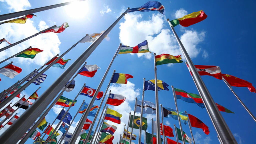 Sur cette photo, plusieurs mâts de drapeaux sont visibles, avec des drapeaux de différentes nations de tous les continents. L'image a été prise d'en bas, en direction du ciel. Les drapeaux flottent tous dans le vent et quelques nuages sont visibles dans le ciel.