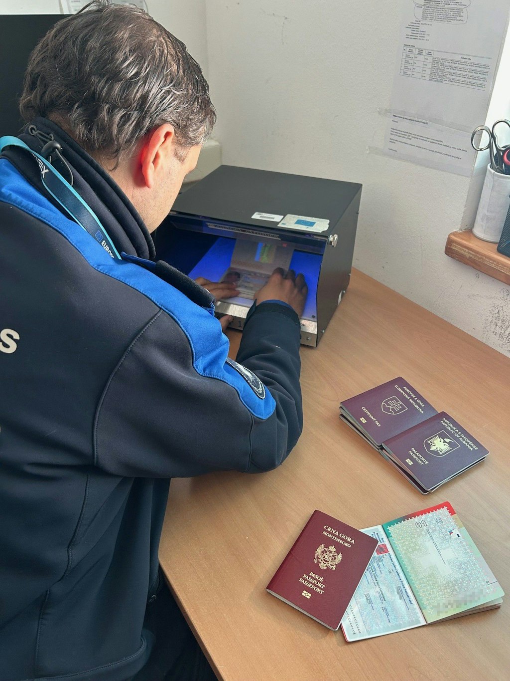 Un agent des douanes verifie un passeport dans un appareil de controle de documents sous lumiere bleue, tandis que d autres passeports sont poses sur le bureau. La scene montre une situation de controle frontalier dans un bureau.  