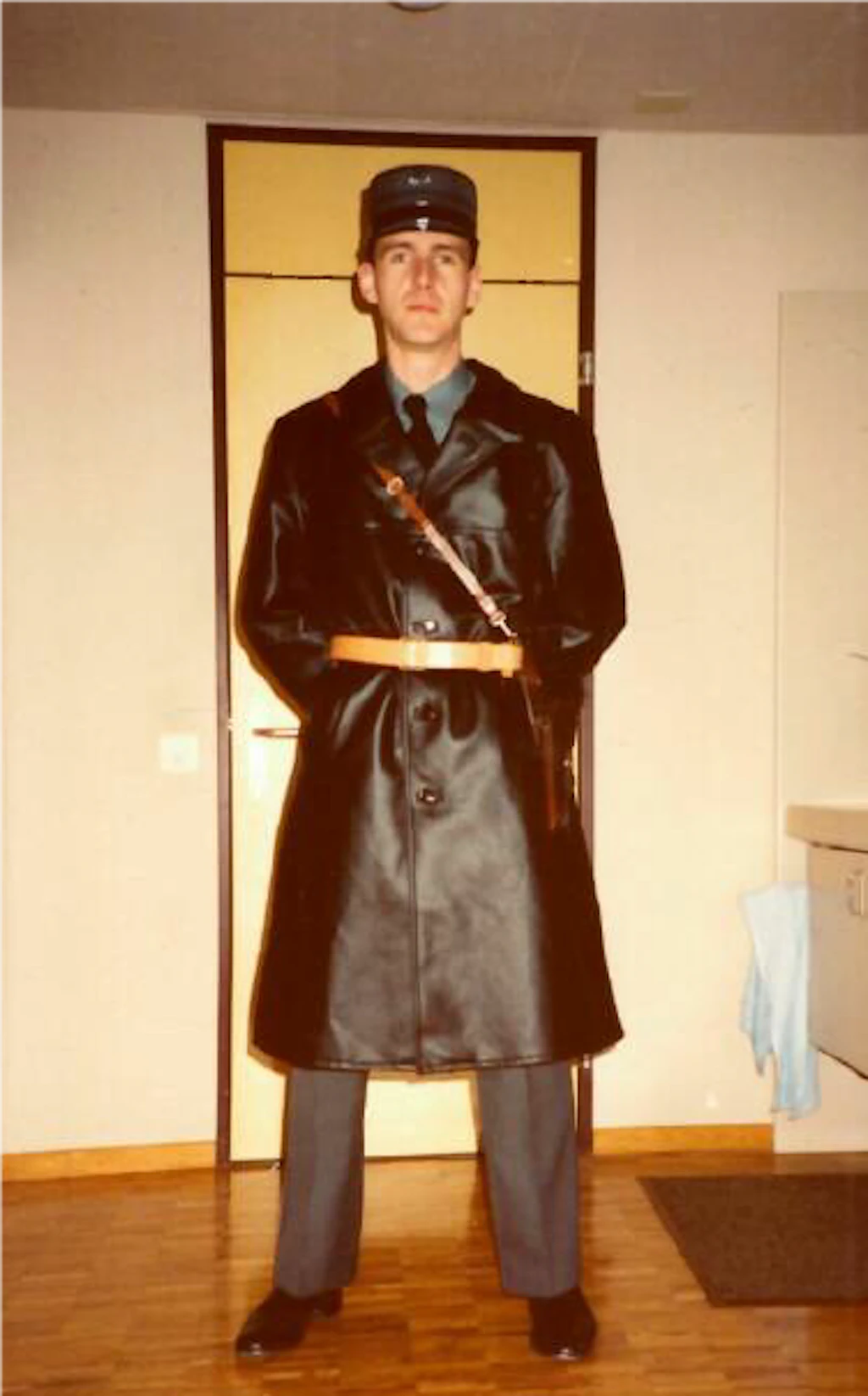 Un jeune aspirant de la garde-frontière se tient dans une piece intérieure, porte un long manteau d'uniforme en similicuir sombre, un chapeau rigide et une bandoulière en cuir. Il regarde droit vers l'objectif, les mains dans les poches du manteau, avec une porte et un coin d'ameublement en arrière-plan.