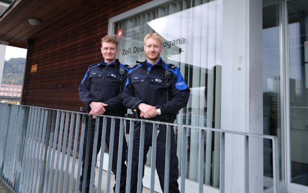 Deux douaniers se tiennent devant un bâtiment de bureaux avec l'inscription "Zoll Douane Dogana". Ils portent des uniformes et sourient à la caméra.