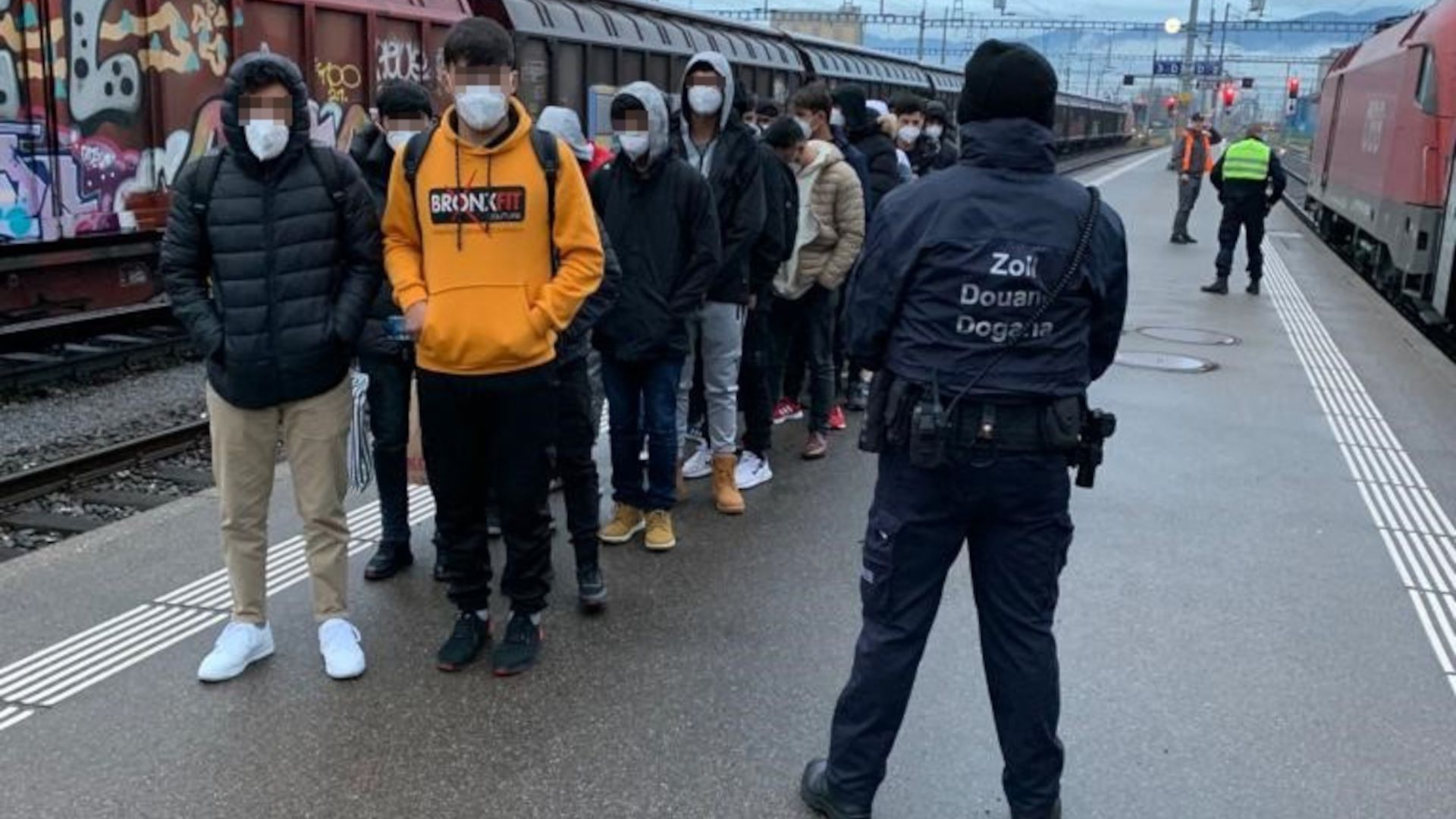 Plusieurs jeunes hommes se tiennent en colonne par deux. Les hommes portent tous une protection bucco-nasale et leurs yeux sont pixélisés. Au premier plan se trouve une personne portant un uniforme bleu foncé des douanes et une casquette noire. Son visage n'est pas visible.