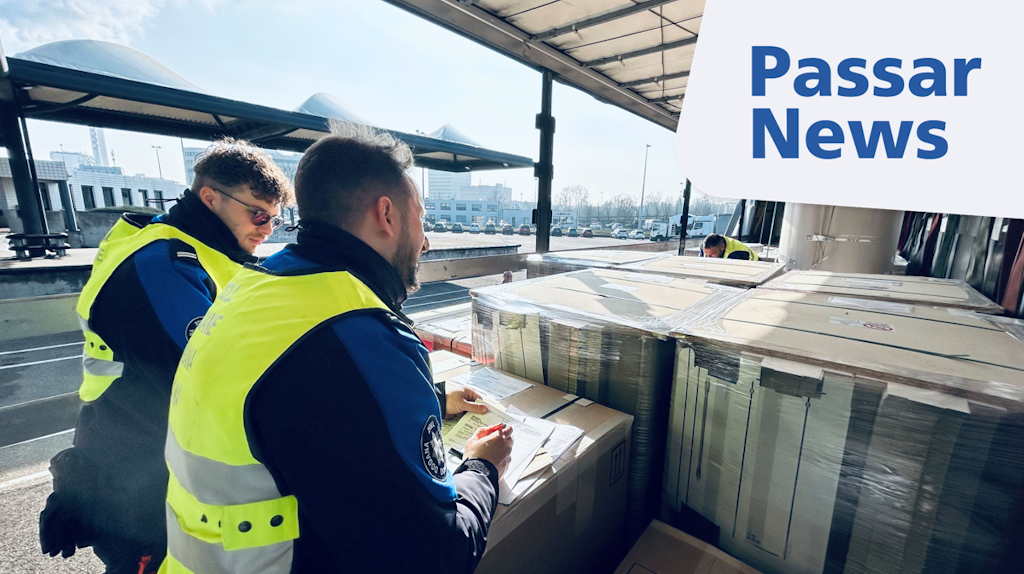 Deux agents des douanes inspectent des palettes chargées et vérifient les documents correspondants à un poste de contrôle frontalier. En arrière-plan, on distingue les installations extérieures d’un site moderne.