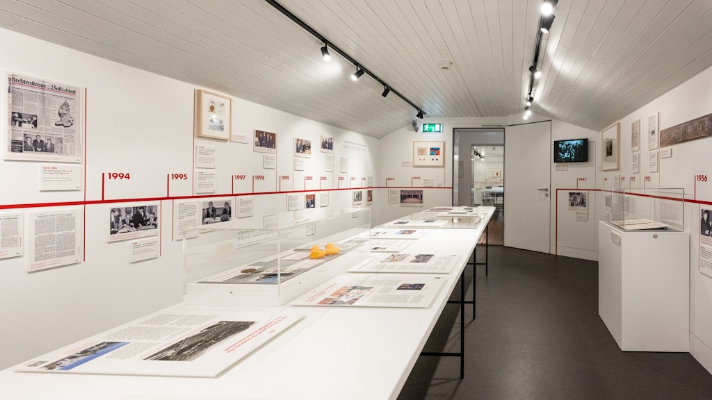 Vue d’une salle d’exposition lumineuse du musée des douanes avec une longue vitrine centrale et une frise chronologique rouge sur le mur. Des documents historiques, des photos et des objets illustrent l’histoire des douanes.