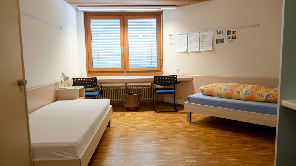 Une chambre double simplement meublée avec deux lits simples, deux chaises, une fenêtre avec des stores et un sol en bois. Des feuilles d'information sont accrochées au mur et une couette colorée est posée sur un lit. La chambre paraît propre et fonctionnelle.