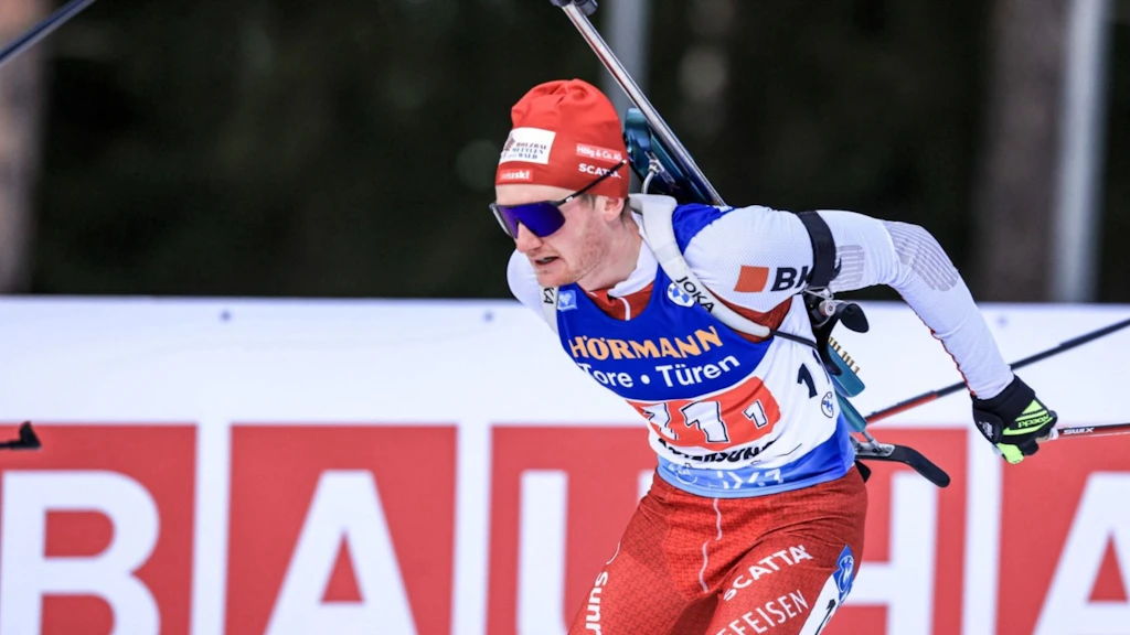 Un biathlète en tenue rouge de compétition et dossard bleu court avec des skis et une carabine sur le dos. À l’arrière-plan, on voit une affiche publicitaire. La scène montre une compétition sportive en plein air.  