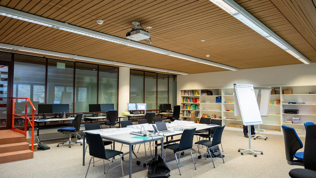 Une salle de réunion lumineuse avec plusieurs tables, chaises, ordinateurs et tableaux à feuilles. À l’arrière-plan, des étagères contiennent des livres et du matériel de travail. L’ambiance est professionnelle et organisée.
