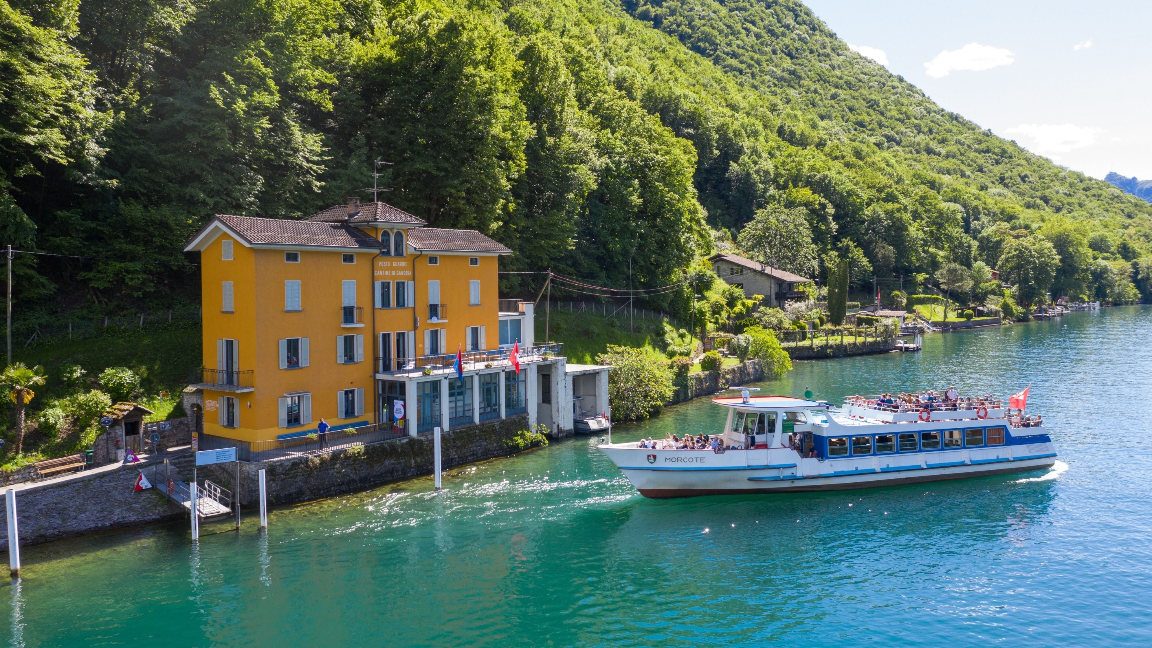 Un bâtiment jaune au bord du lac avec un bateau de croisière devant, entouré de verdure.