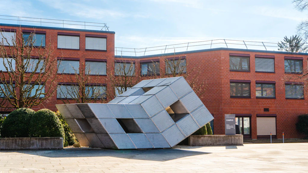 Une sculpture métallique géométrique moderne se trouve sur une place devant un bâtiment en briques rouges avec de nombreuses fenêtres. À l'arrière-plan, on voit des arbres taillés et un panneau indiquant "Campus". La scène est calme et ensoleillée.