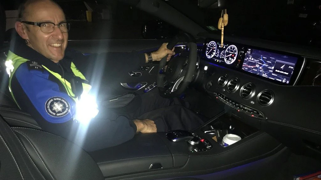 Un agent en uniforme du service des douanes et de la sécurité des frontières est assis au volant d’un véhicule de patrouille moderne de nuit, avec le tableau de bord éclaire. Il sourit vers l’appareil photo tandis que les écrans de navigation et de vitesse brillent en arrière-plan.