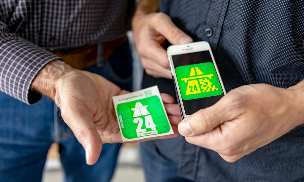 Deux personnes tiennent la vignette autoroutiere suisse 2024, une fois comme vignette autocollante et une fois comme e-vignette sur un smartphone. Les vignettes vertes montrent le symbole de l autoroute et l annee 24.  