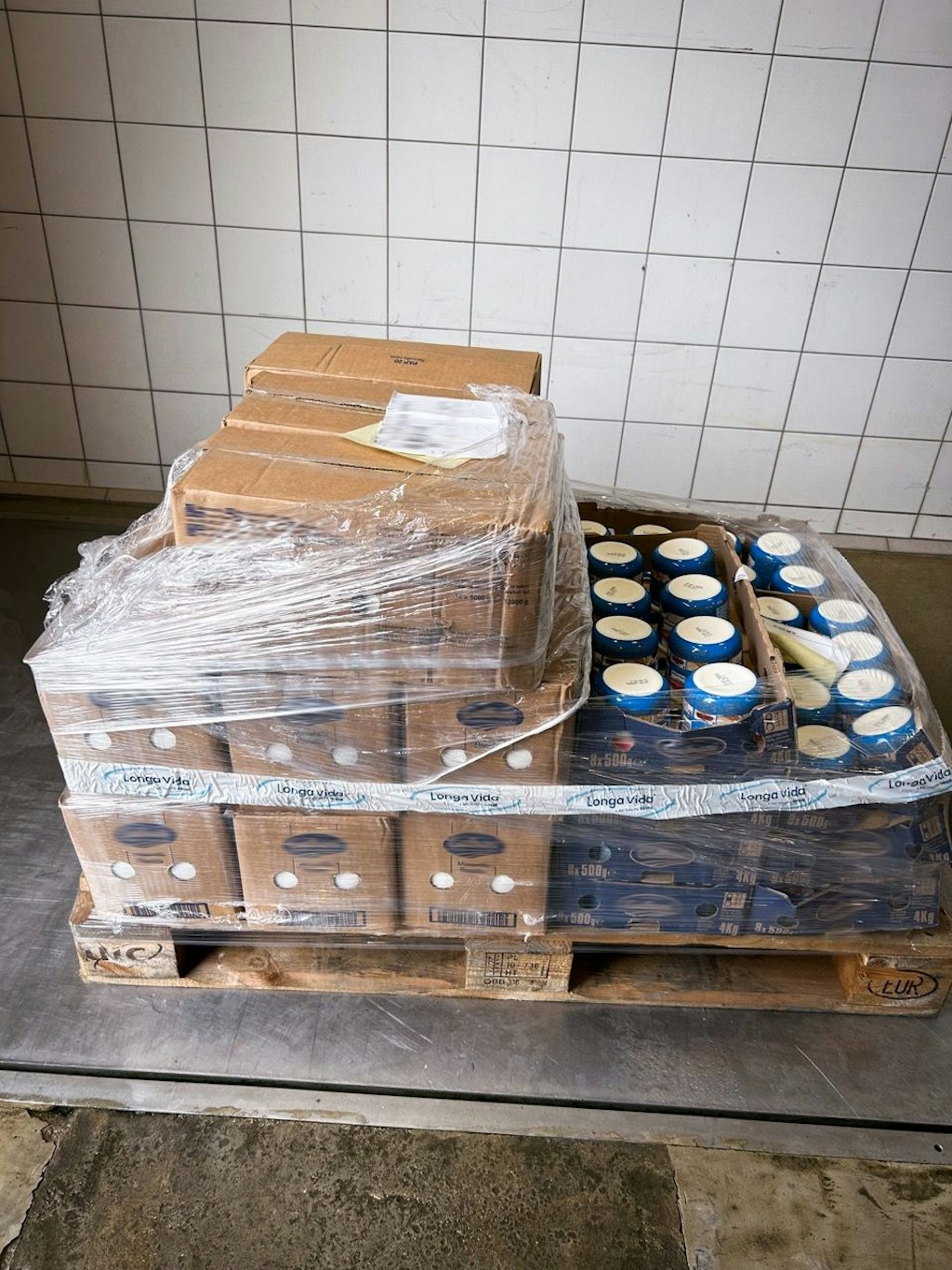 Une palette avec divers aliments, y compris des boîtes et des contenants enveloppés dans du plastique. Les marchandises comprennent des produits comme la burrata, la mozzarella, l'édam et les jaunes d'œufs.