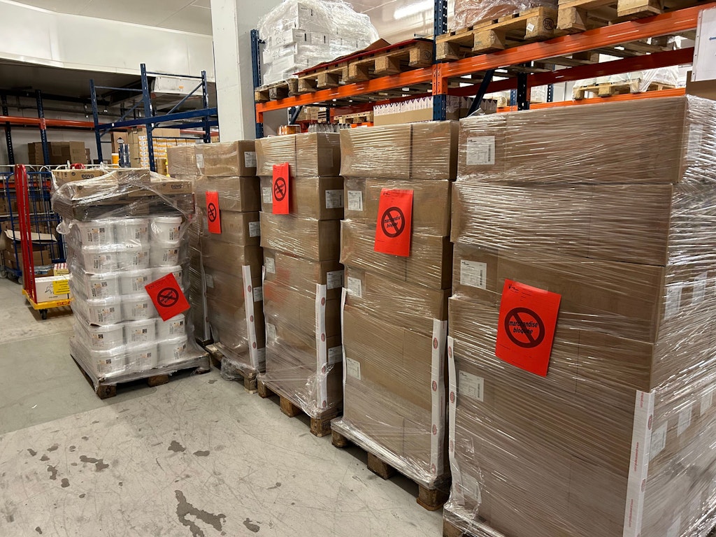 Palettes avec des cartons et des panneaux rouges indiquant une saisie. Les marchandises sont empilées dans un entrepôt.