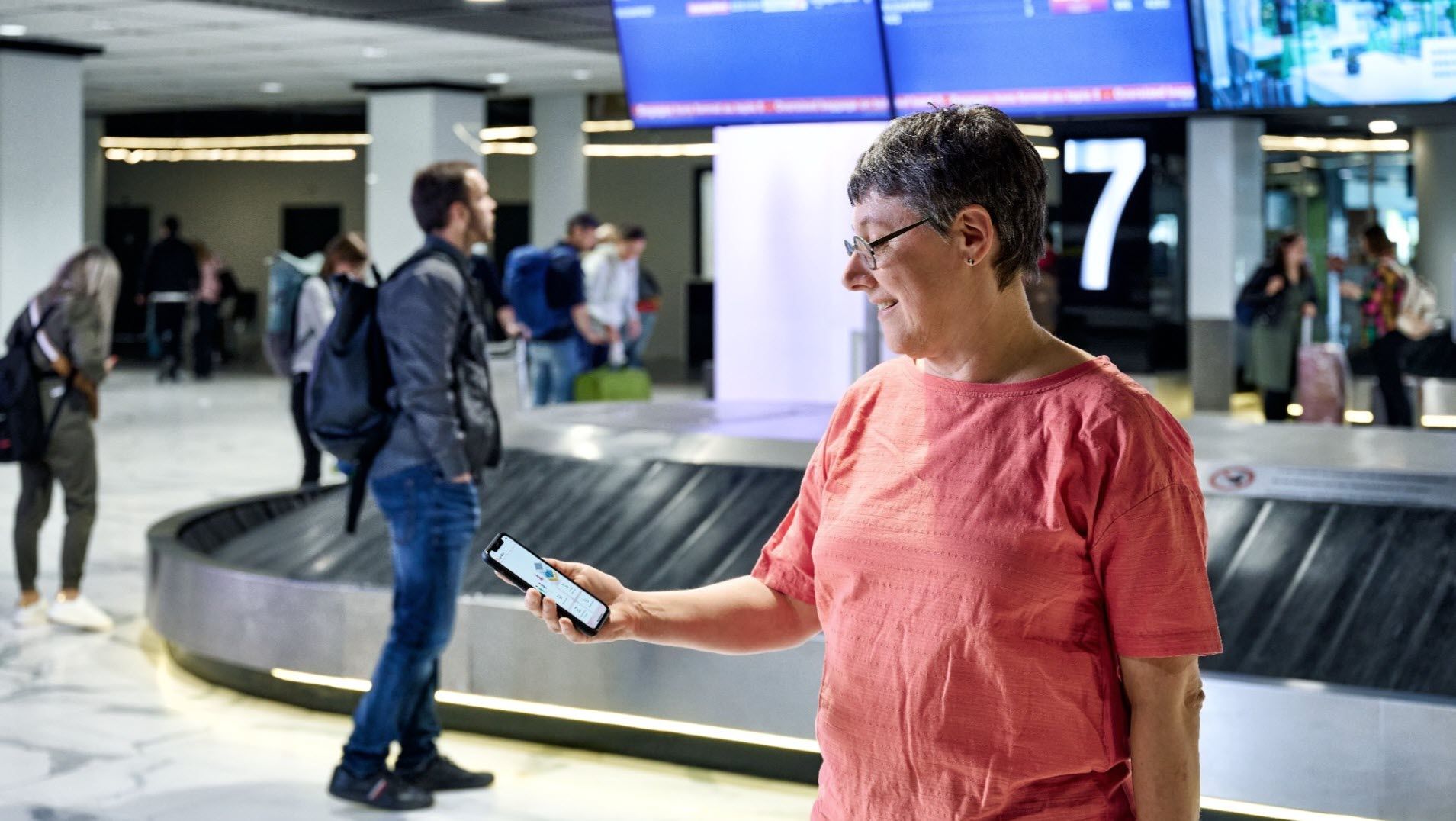 Une femme se tient près d'un tapis à bagages à l'aéroport et regarde son smartphone. D'autres voyageurs sont visibles en arrière-plan. 