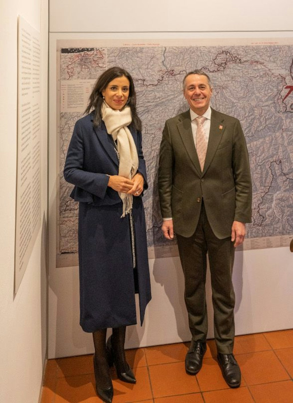 Deux personnes élégamment habillées se tiennent côte à côte devant une grande carte historique dans une salle d’exposition. Elles regardent l’objectif et posent pour une photo officielle.