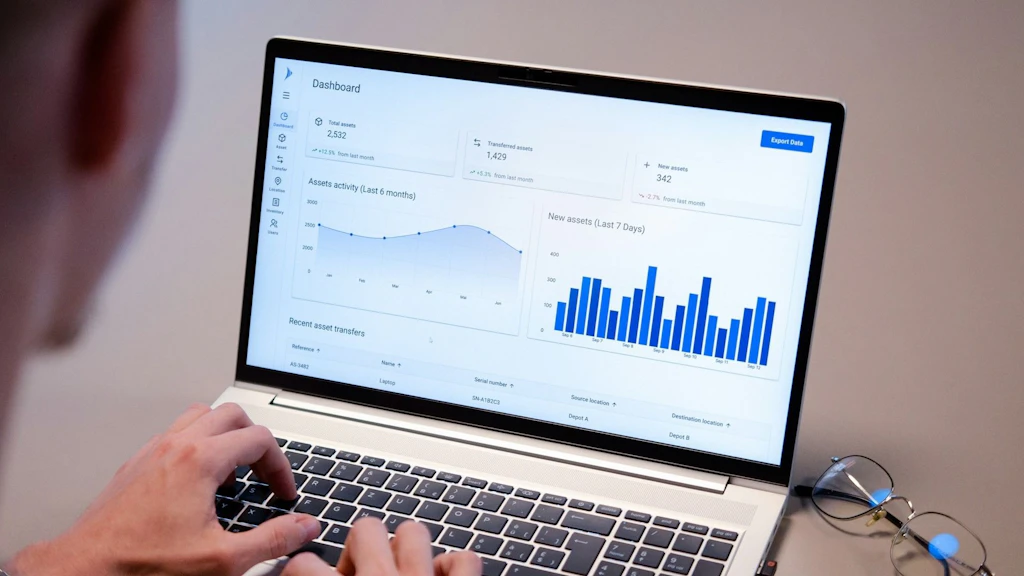 Un ordinateur portable affiche un tableau de bord avec des graphiques et des statistiques. Une personne tape sur le clavier, avec des lunettes à côté.