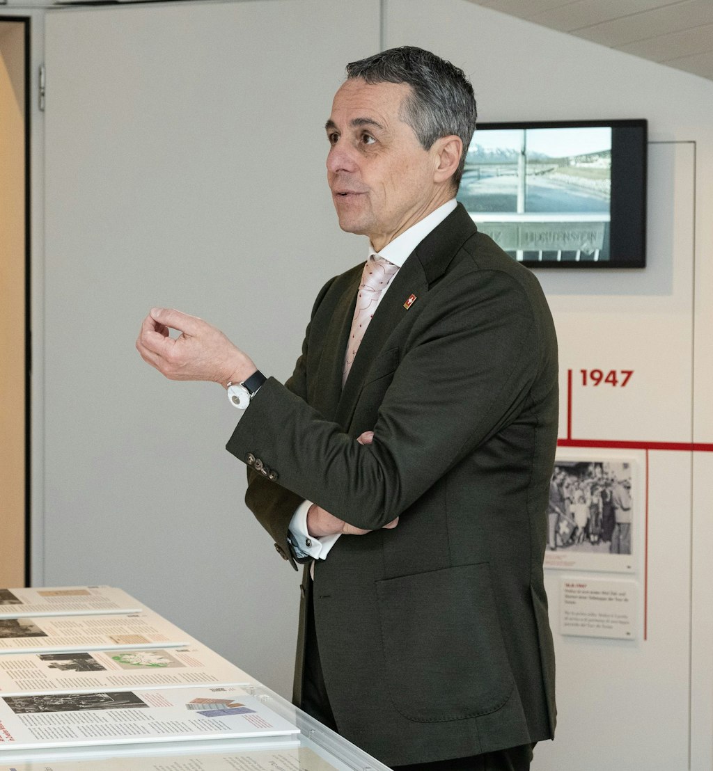 Un homme en costume se tient dans une exposition devant une vitrine avec des panneaux d’information et parle en gesticulant. En arrière-plan, on voit des panneaux muraux avec des textes et un écran.