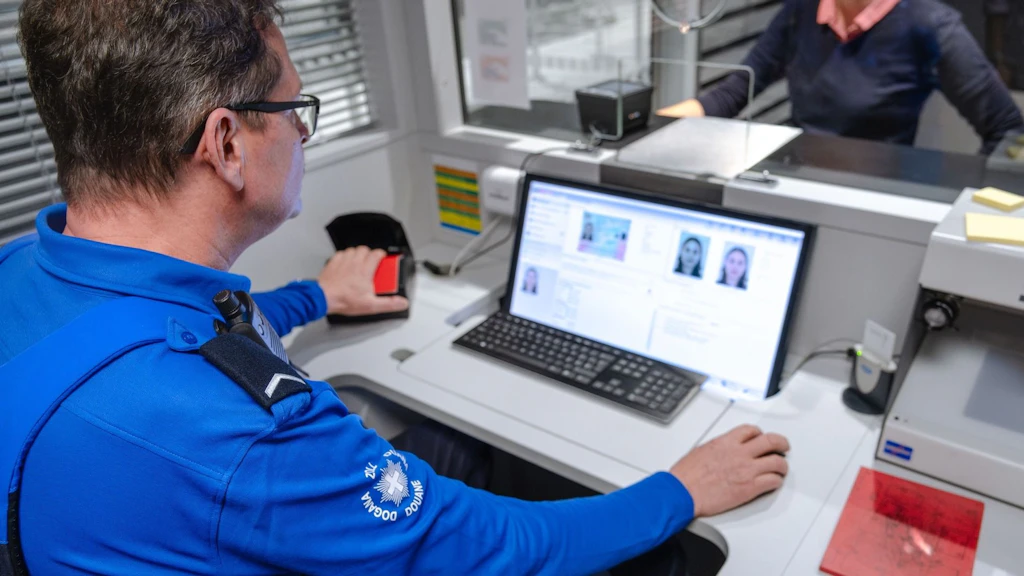 Un collaborateur de la douane vérifie les documents d'identité d'une personne à un guichet à l'aide d'un ordinateur et d'un scanner d'empreintes digitales. En arrière-plan, une personne est assise de l'autre côté de la vitre. La scène illustre une situation typique de contrôle aux frontières.